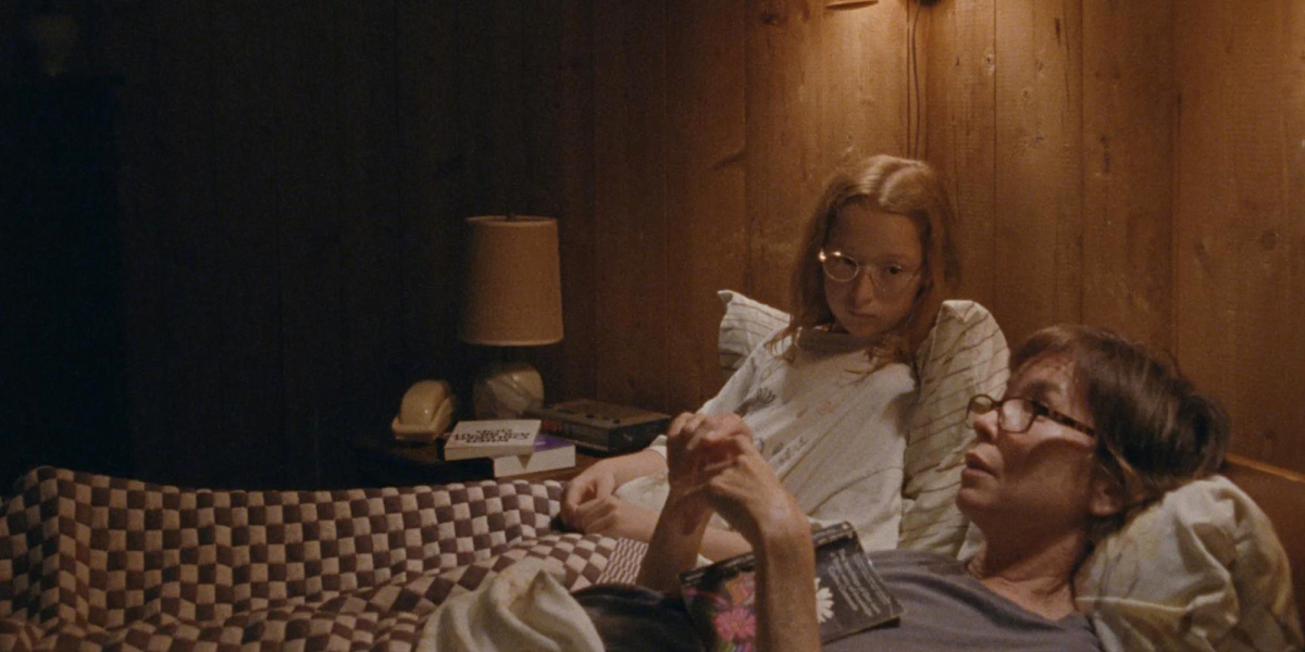 A girl lies in bed next to her mom, both wearing glasses in Janet Planet.