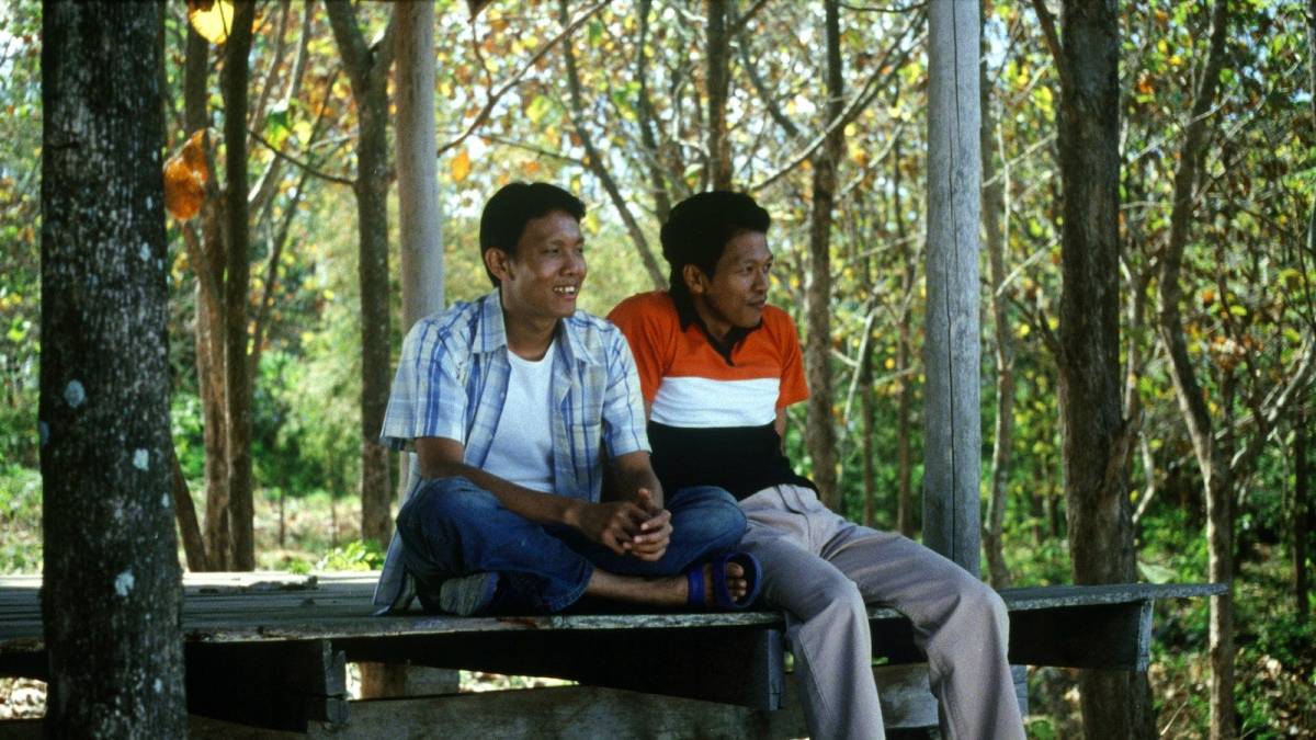 Two men sit next to each other on a dock with trees behind them.