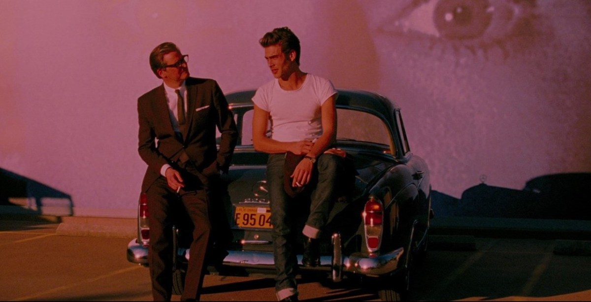 A man in a suit and a man who looks like James Dean lean against a car in pink light with the image of an eye behind them.