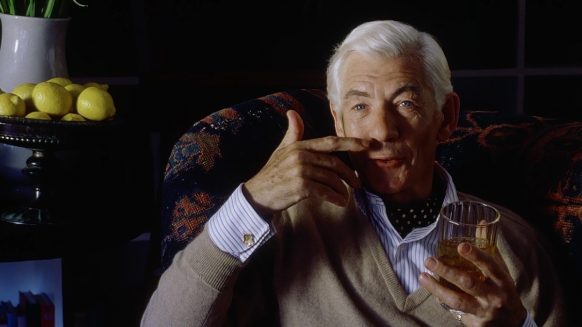 Ian McKellan smiles and points at his upper lip.