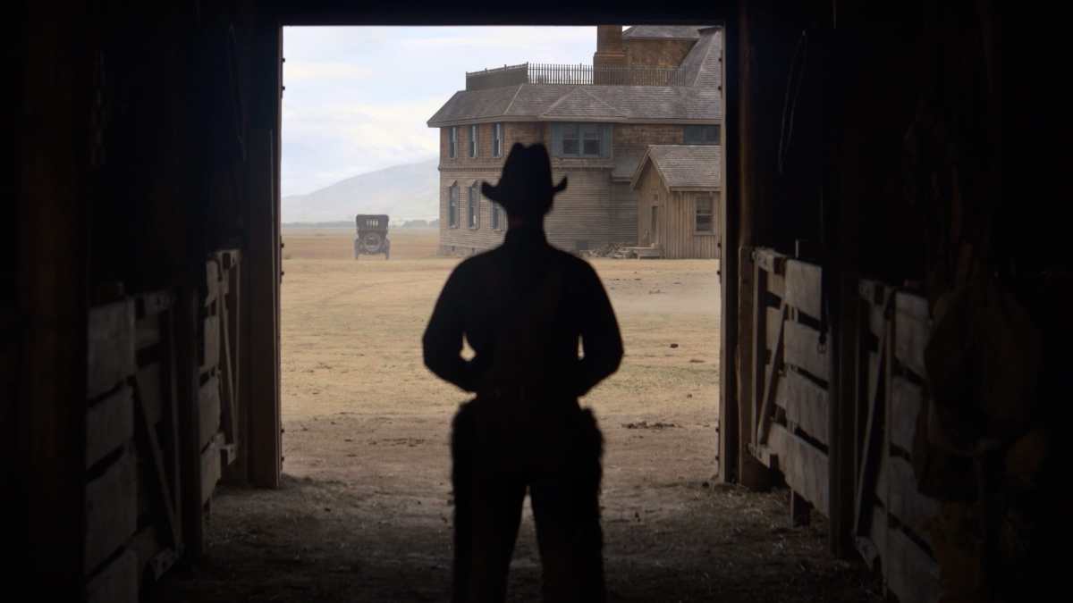 A cowboy stands backlit in the door of a barn.