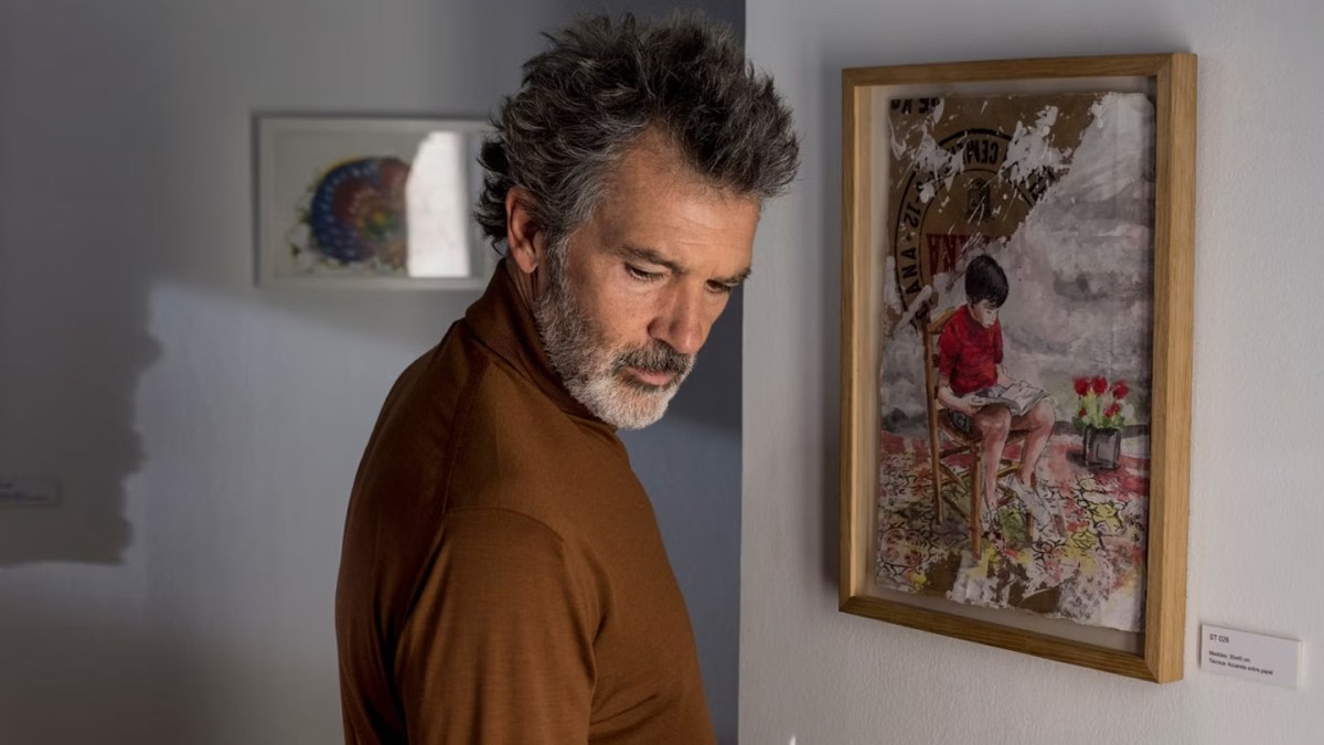 Antonio Banderas with a hair style like Almodóvar's looks over his shoulder with a painting of a boy on the wall behind him.