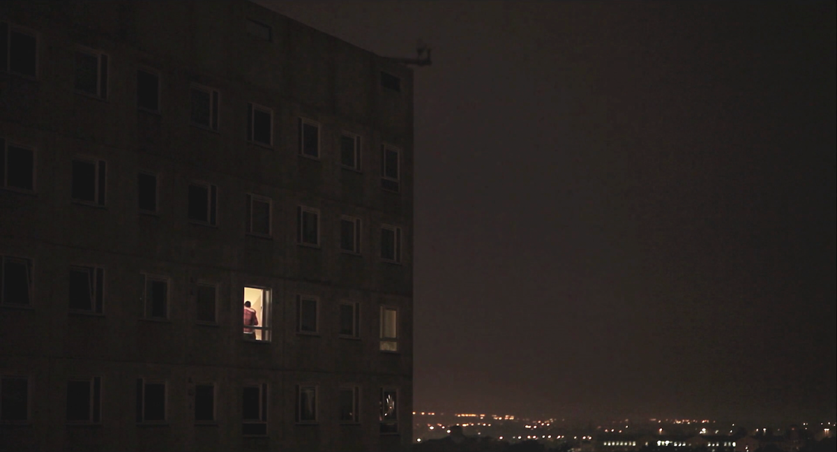 A wide exterior shot of an apartment complex. Only one apartment has the light on and the figure of two people is visible.