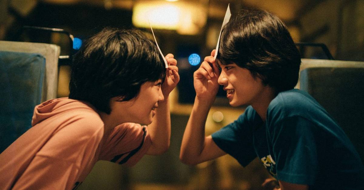 Two young boys look at each other as they play a game where they hold cards up to their foreheads.