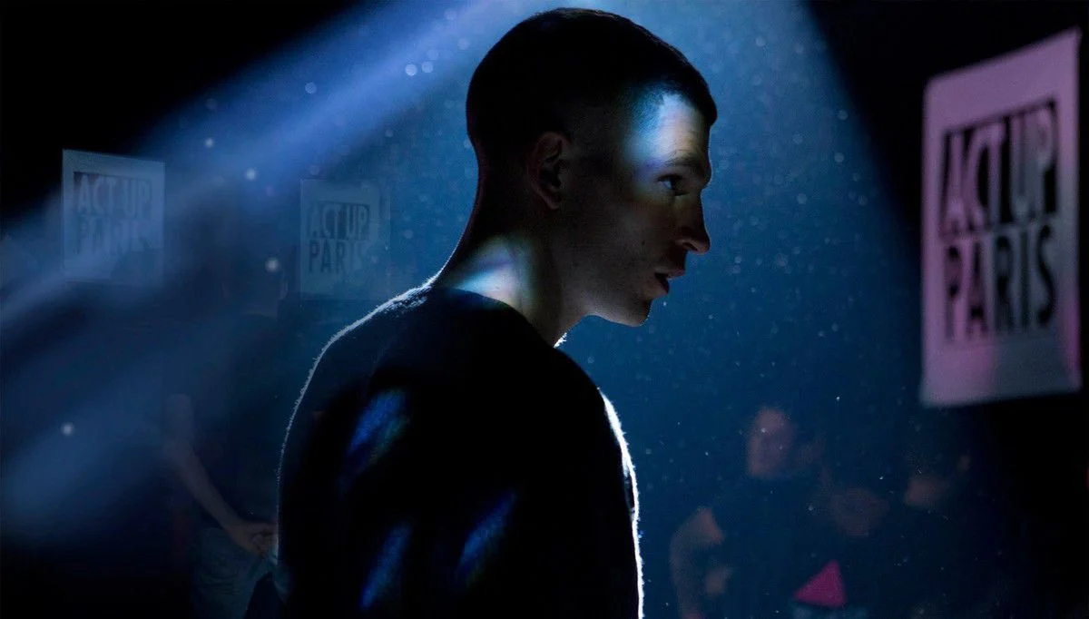 A gay man stands in a spotlight with ACT UP Paris posters in the background.