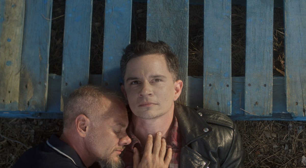 Two trans men cuddle, heads resting on wooden planks as one looks up to the sky.