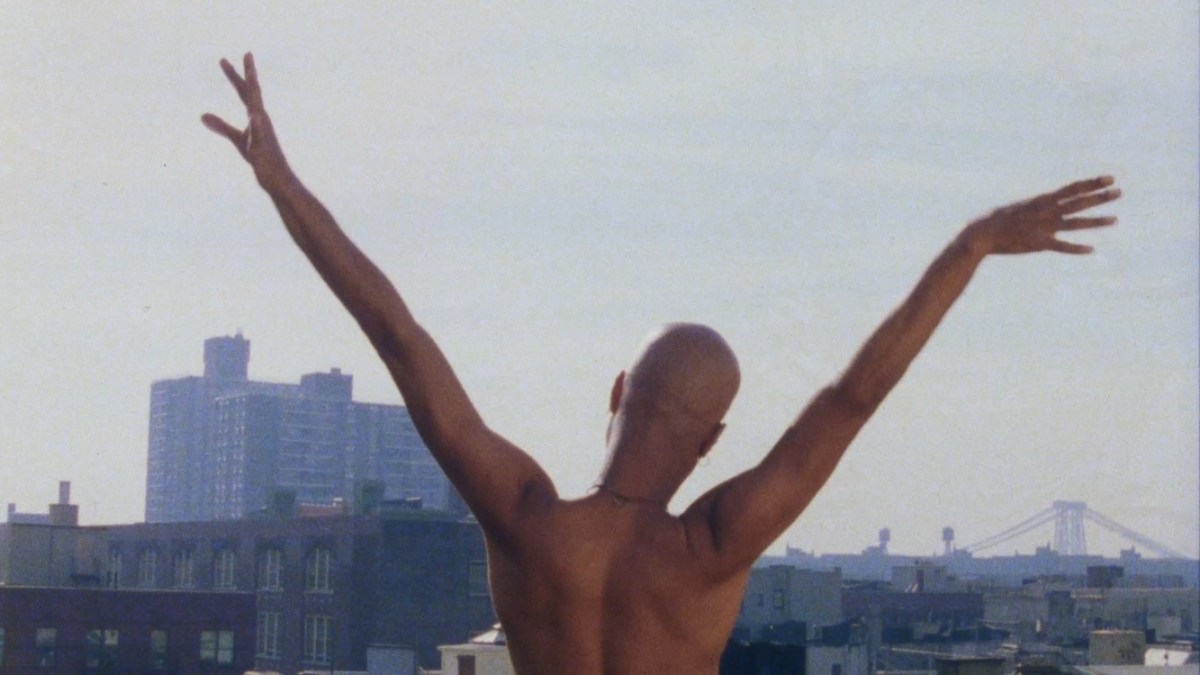 A bald Black man lifts his arms in the air while looking out over a cityscape.