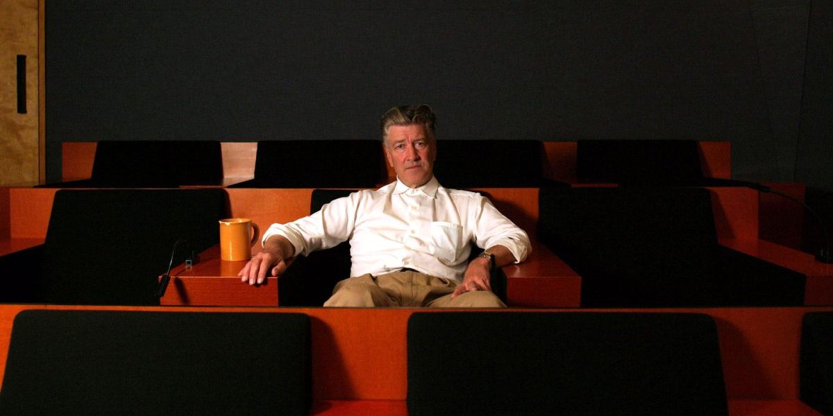 David Lynch sits in a screening room alone on a red seat looking at the camera.