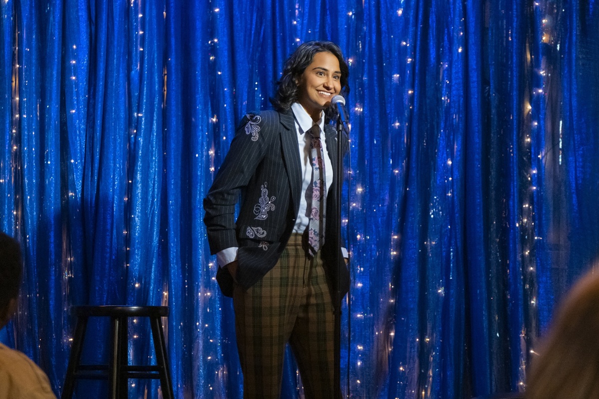 Standing on stage, in front of a blue curtain with sparkles, Bela approaches the microphone to participate in the storytelling event. She's wearing a bunch of suit separates: brown plaid pants, light blue shirt, tie and blue blazer.