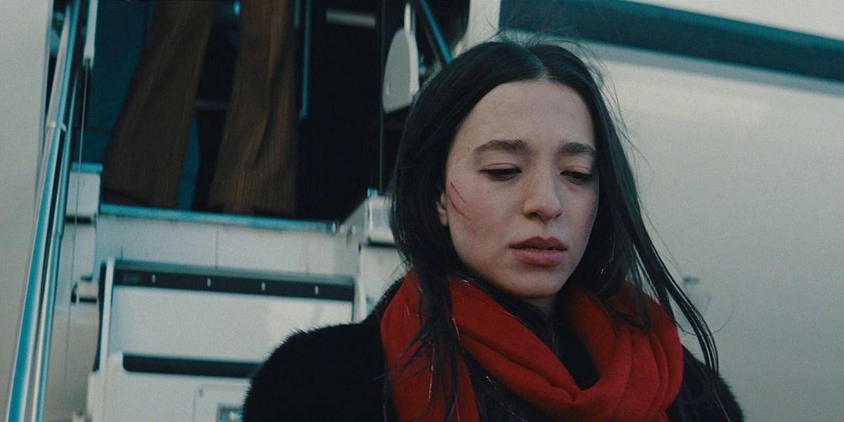 Mikey Madison with a scratched face and a red scarf looks down standing in front of an airplane staircase.