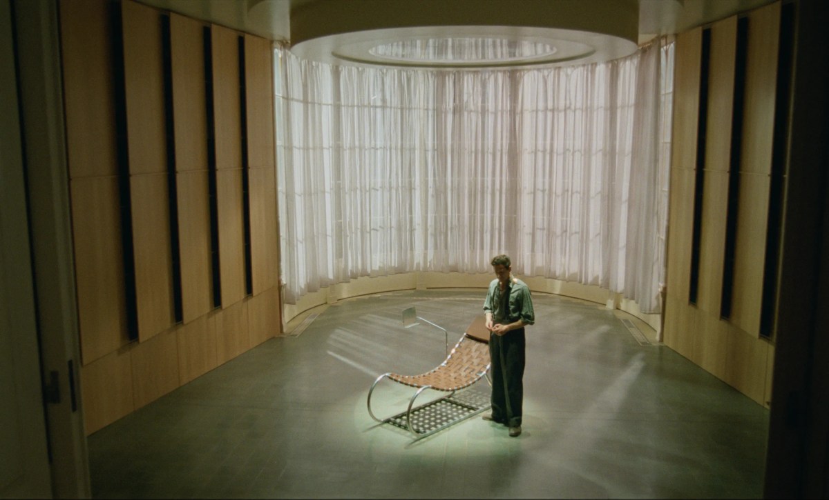 Adrien Brody stands in the middle of a brutalist library with a single chair in the middle