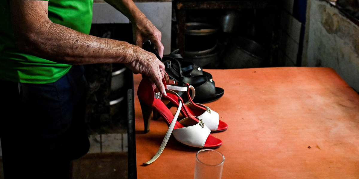 The arms of an older person displays two pairs of high heels on a wooden table.